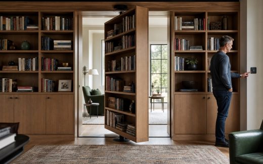 Motorized rotating oak bookcase storage wall partially pivoted open in a contemporary home library with a person operating a wall control