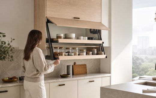 Contemporary light-oak and warm-white kitchen with a motorized lift-down wall cabinet lowered to bring shelves of plates and pantry jars within reach